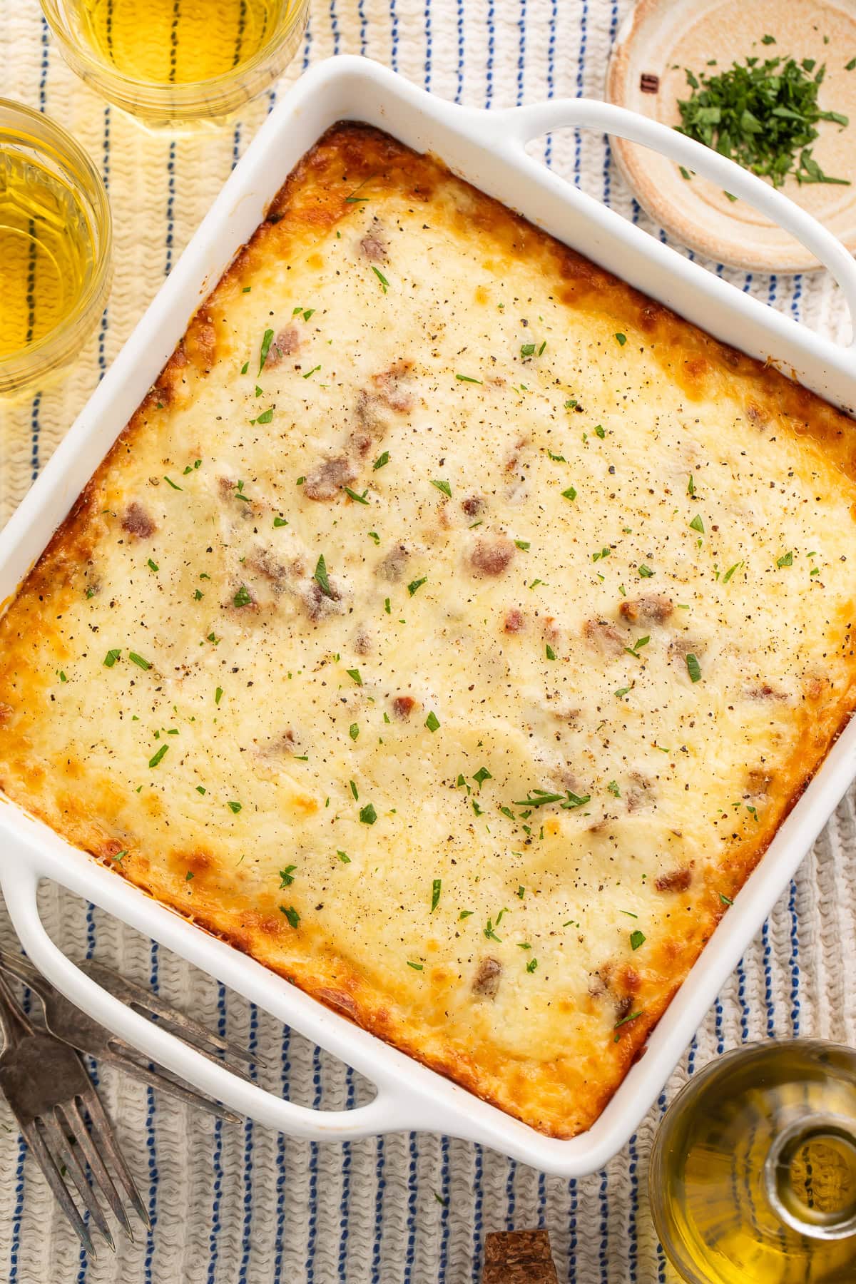 White dish of baked white lasagna with a bowl of parsley, bottle of olive oil and cups of beer visible just off to the side.