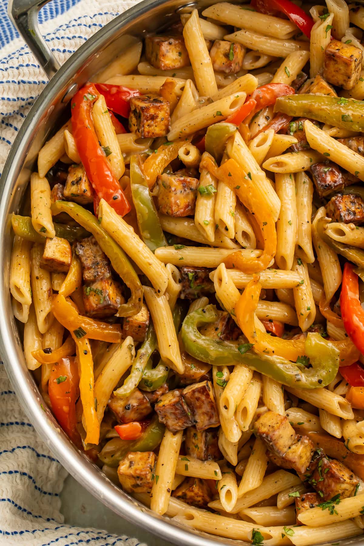 Pan of vegan rasta pasta with bell peppers and tofu on top of a blue and white kitchen towel, pictured in close up.