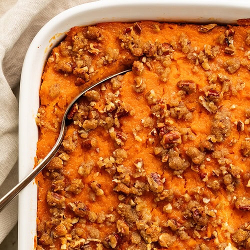 Close up of a sweet potato casserole topped with pecans in a white baking dish. A spoon has been inserted in the corner to lift out a portion.