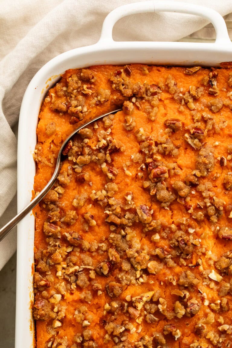 Close up of a sweet potato casserole topped with pecans in a white baking dish. A spoon has been inserted in the corner to lift out a portion.