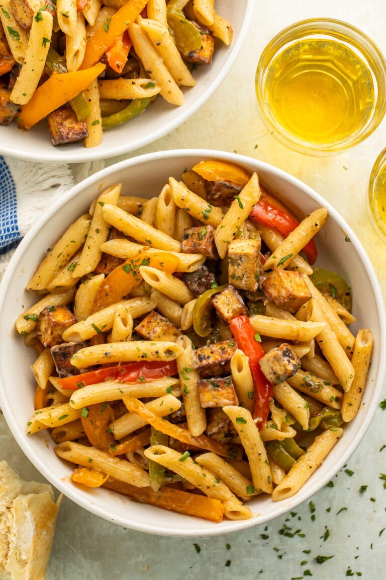 Two white bowls of vegan rasta pasta with glasses of wine pictured off to the side.