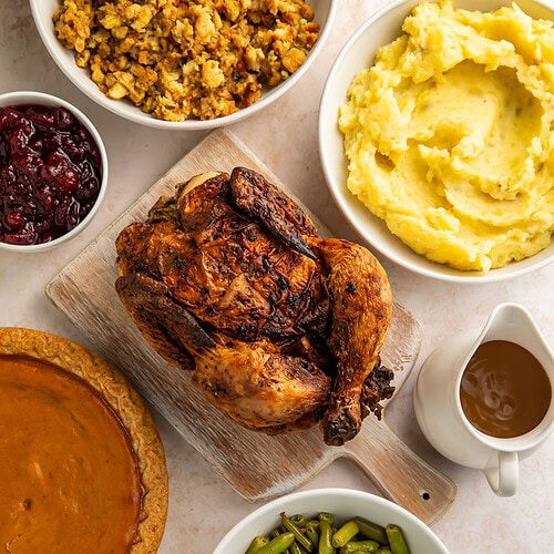 Roast chicken surrounded by holiday trimmings of stuffing, mashed potatoes, gravy, green beans, cranberry sauce and a pie.
