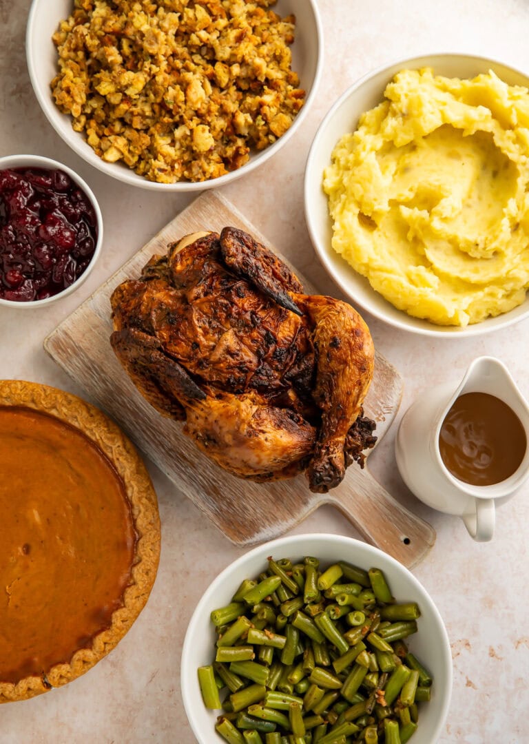 Roast chicken surrounded by holiday trimmings of stuffing, mashed potatoes, gravy, green beans, cranberry sauce and a pie.