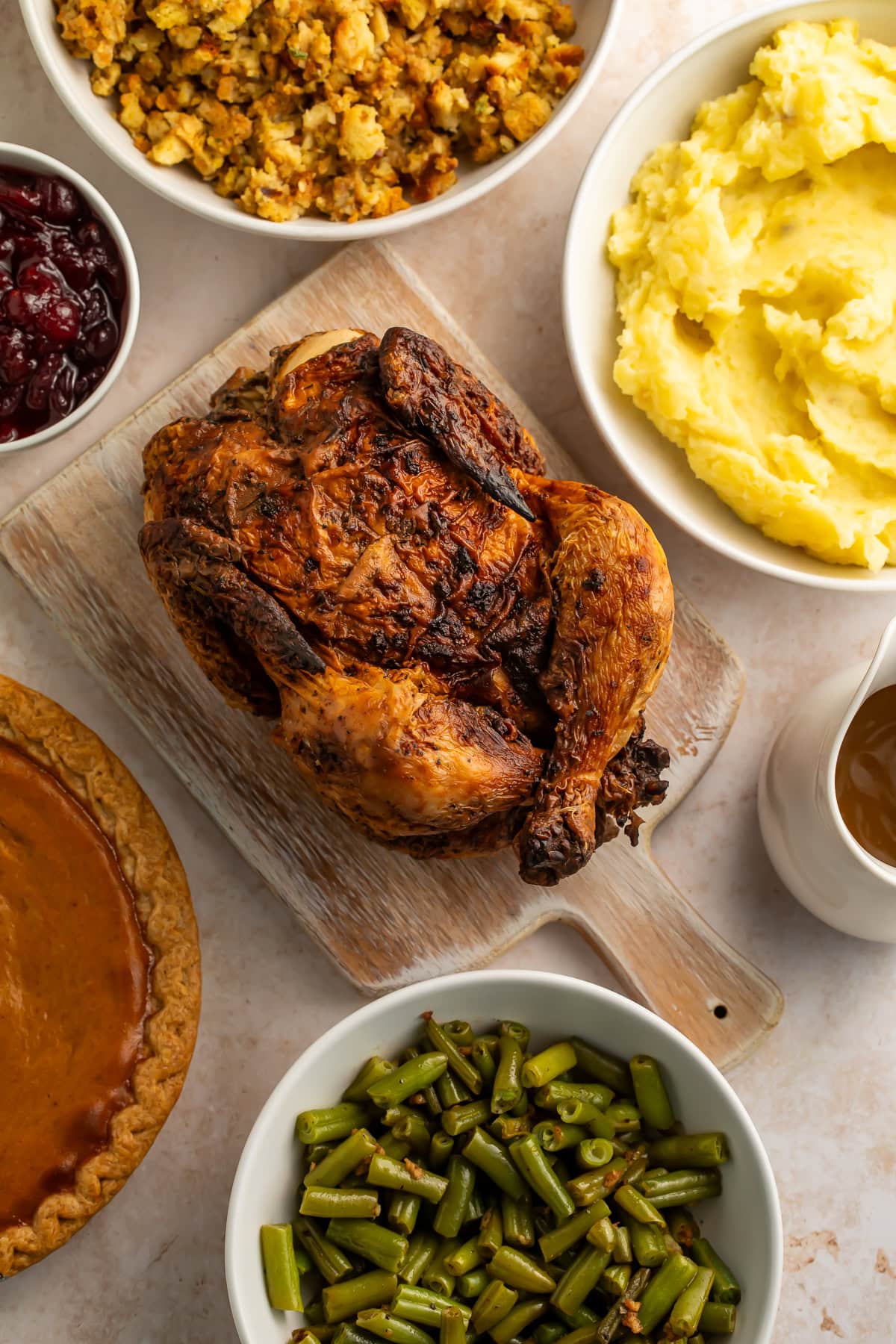 Close up image of Roast chicken surrounded by holiday trimmings of stuffing, mashed potatoes, gravy, green beans, cranberry sauce and a pie.