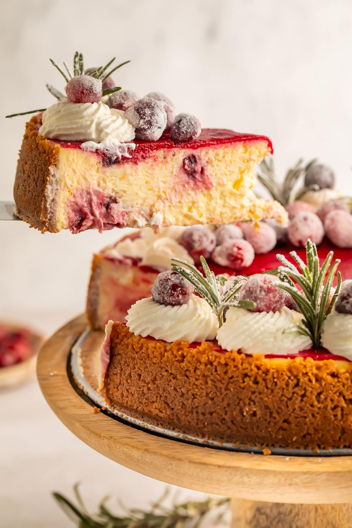Slice of cranberry cheesecake being lifted out of the cheesecake on a wooden cake stand.