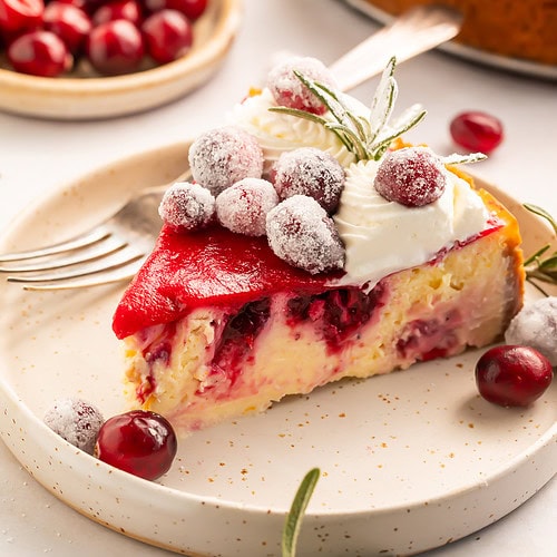 Slice of cranberry cheesecake decorated with whipped cream, sugared cranberries and fresh rosemary sprigs on a white plate.