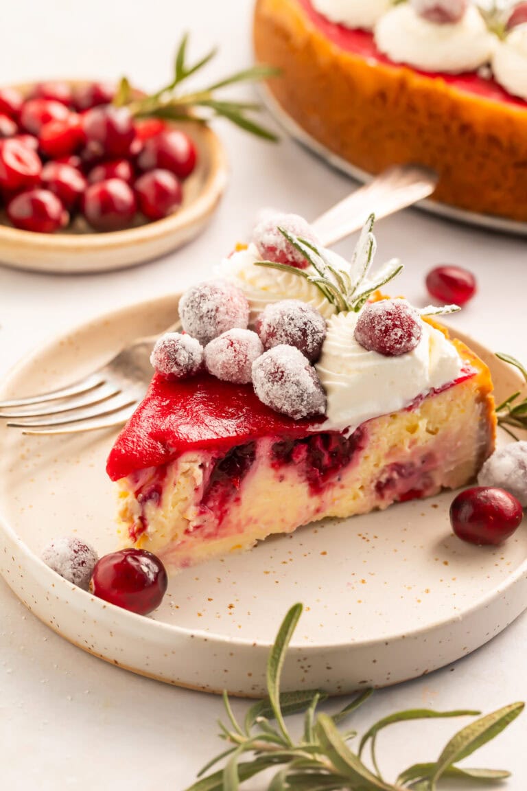 Slice of cranberry cheesecake decorated with whipped cream, sugared cranberries and fresh rosemary sprigs on a white plate.