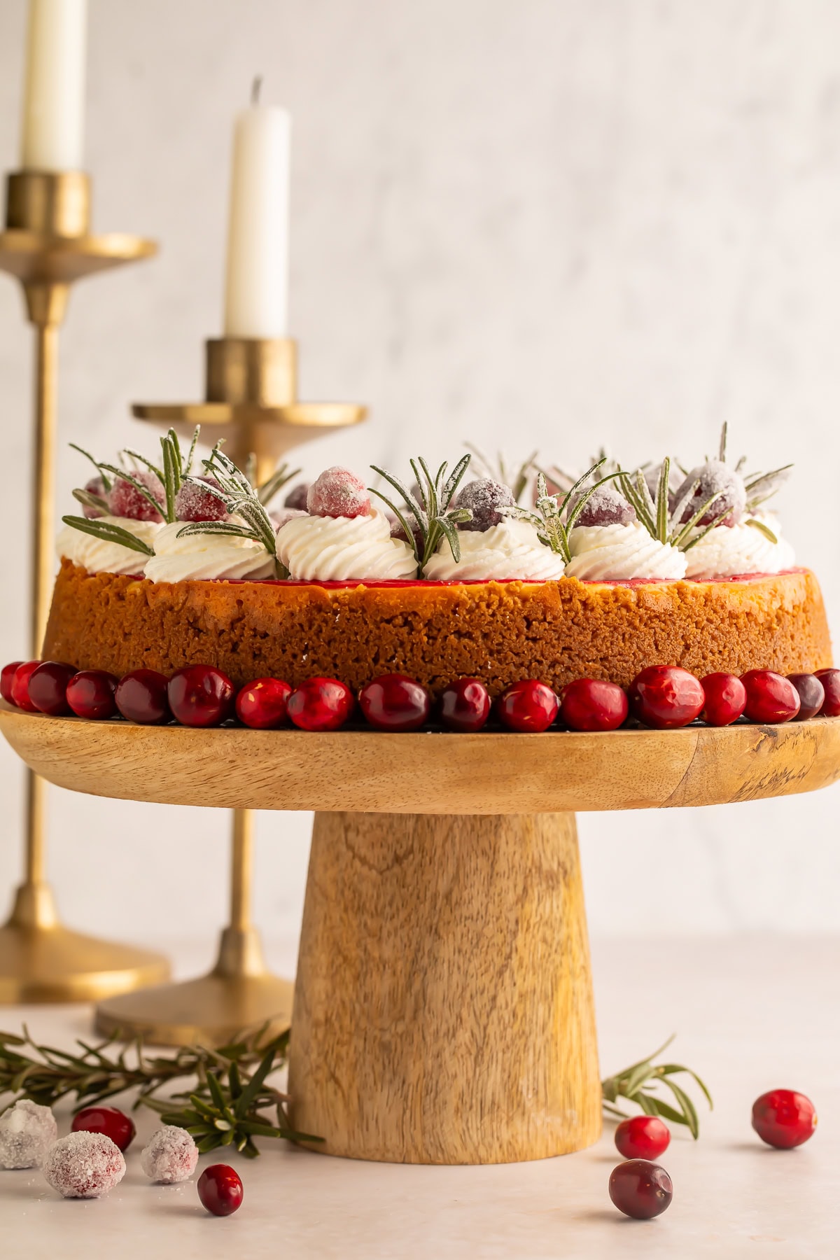 Side on view of a whole cranberry cheesecake decorated with cranberries, cream and rosemary on a wooden cake stand.