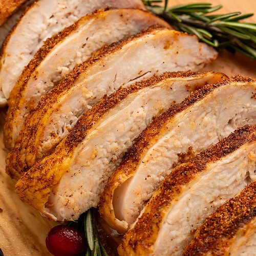 Close up of a sliced turkey breast on a cutting board scattered with rosemary, black grapes and cranberries.