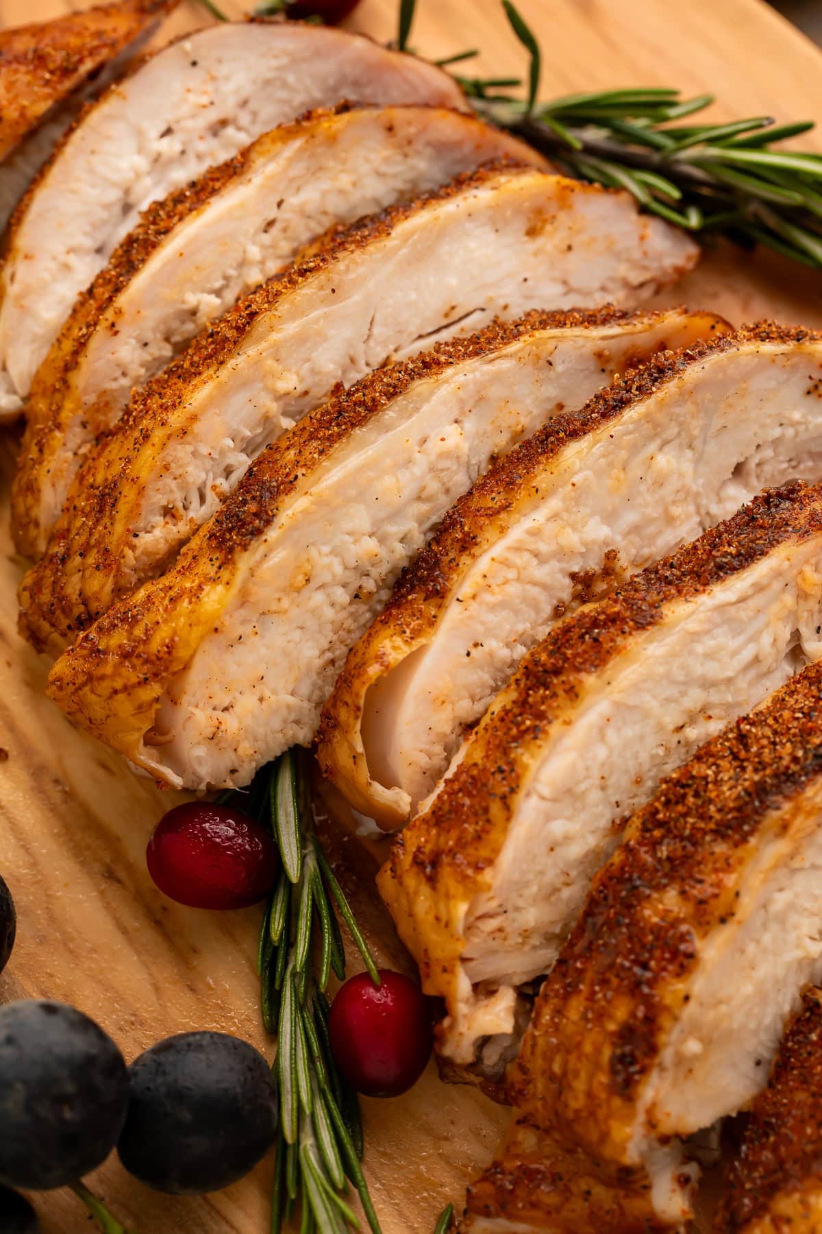 Close up of a sliced turkey breast on a cutting board scattered with rosemary, black grapes and cranberries.