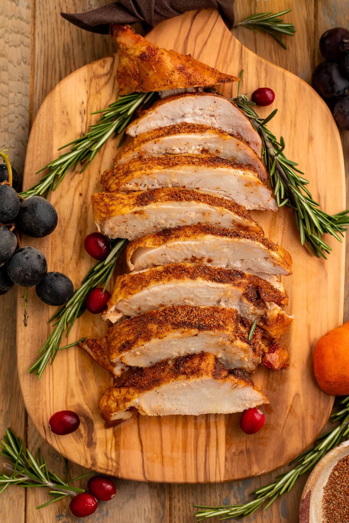 Overhead image of a sliced smoked turkey breast on a cutting board garnished with rosemary sprigs, black grapes, a clementine and cranberries.