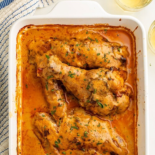 White baking dish of smothered turkey wings pictured top down with a glass of wine and blue and white kitchen towel visible just off to the side.