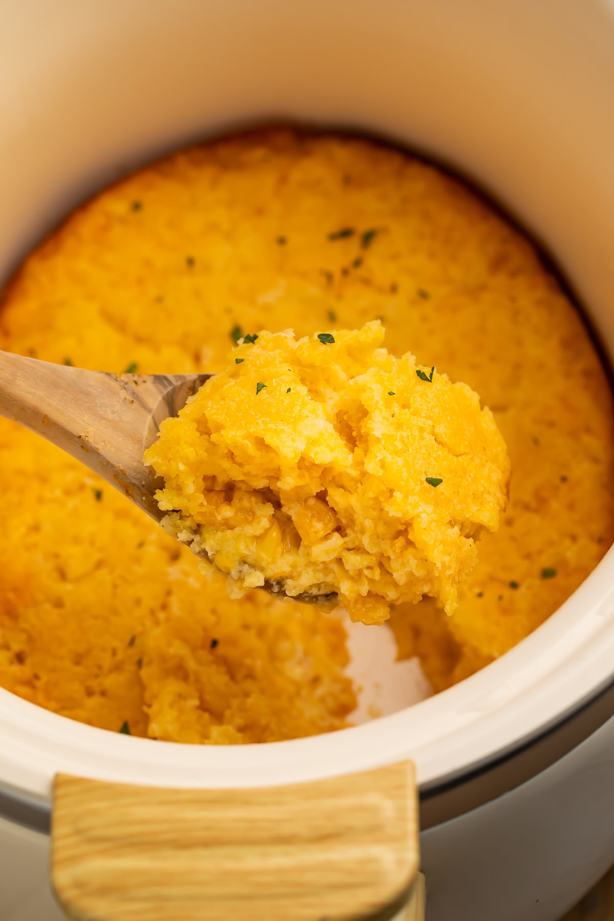 Wooden spoon lifting corn casserole out of the crockpot.