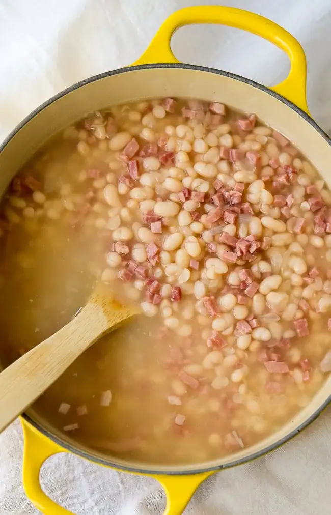 Navy bean and ham soup being stirred in a yellow Dutch oven.