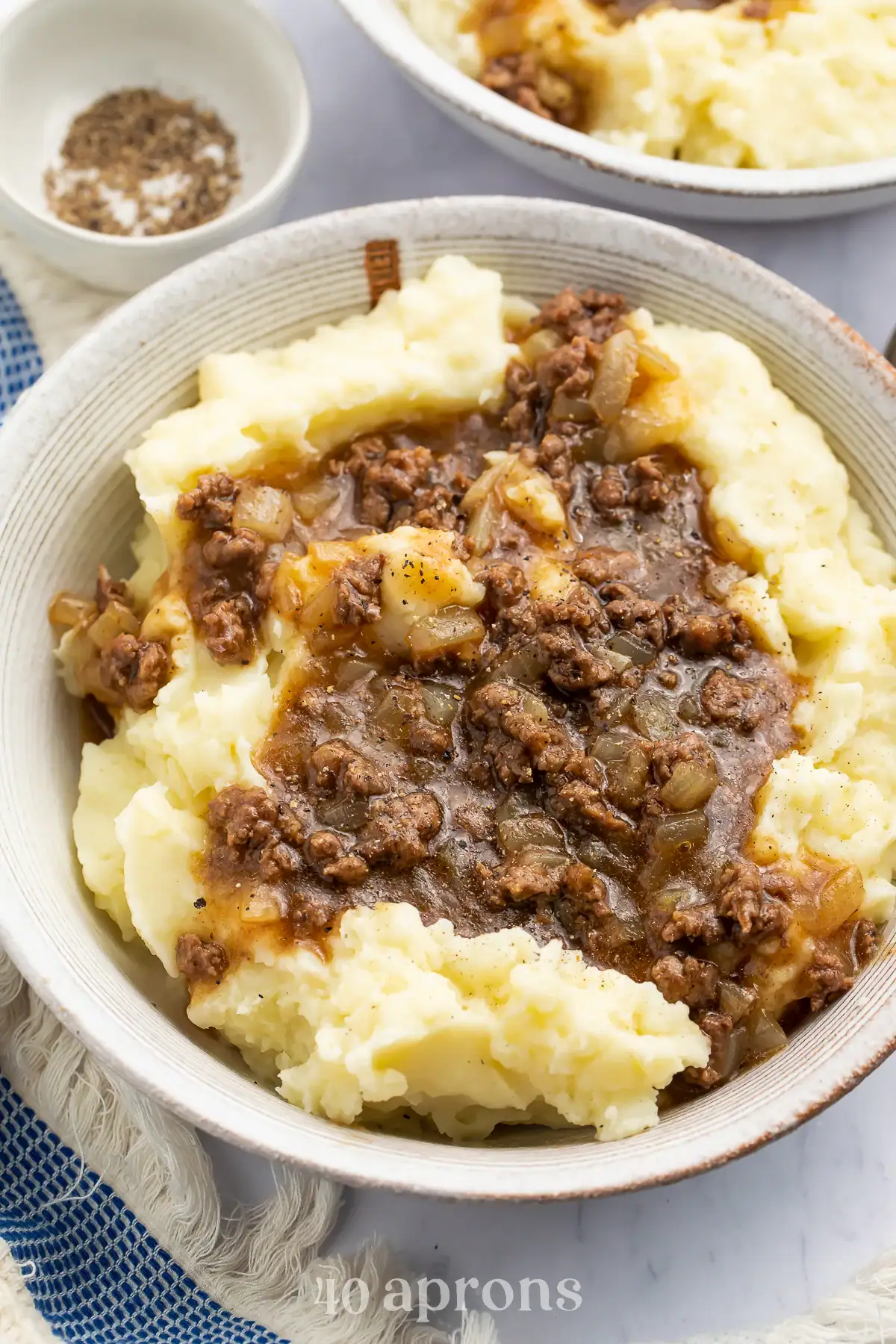 Bowl of mashed potatoes topped with ground beef and gravy. A bowl of black pepper is visible in the background.
