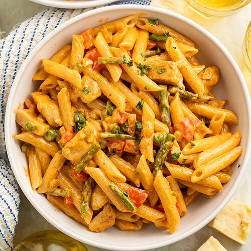 Two white bowls of spicy chicken pasta in a creamy sauce. Glasses of wine and blocks of cheese are off to the side.