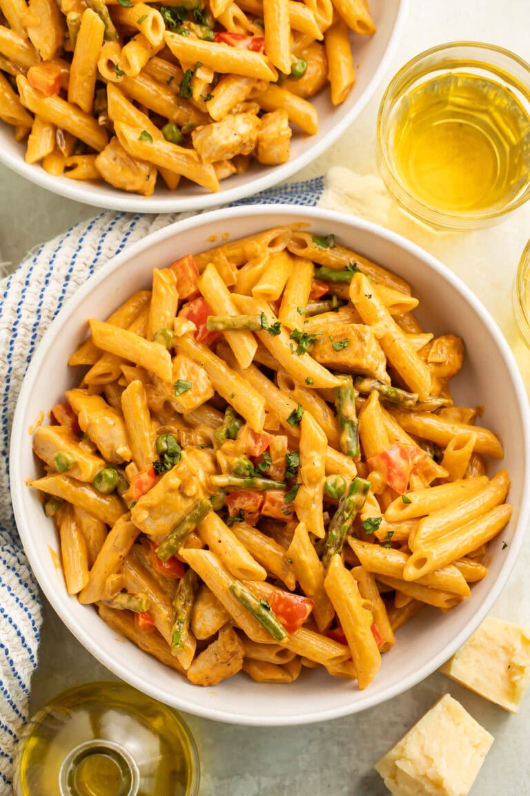 Two white bowls of spicy chicken pasta in a creamy sauce. Glasses of wine and blocks of cheese are off to the side.
