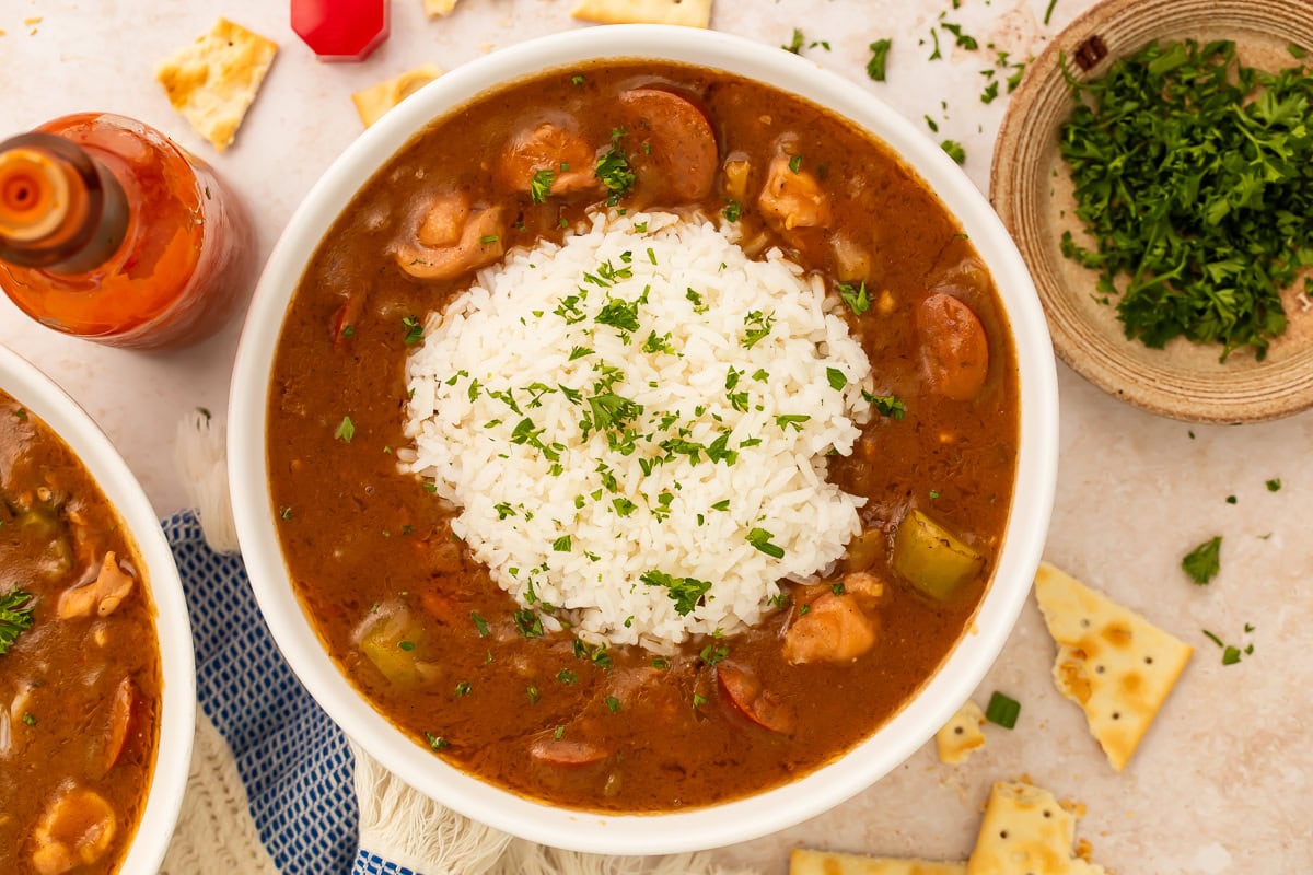 White bowl of gumbo and rice with bowls of herbs, crackers and hot sauce next to it.