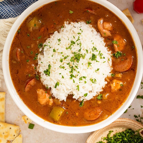 Bowl of chicken sausage gumbo topped with a scoop of rice. Hot sauce and chopped herbs are off to one side.