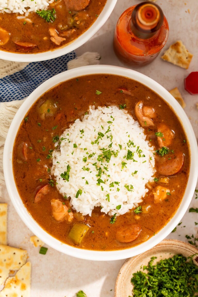 Bowl of chicken sausage gumbo topped with a scoop of rice. Hot sauce and chopped herbs are off to one side.