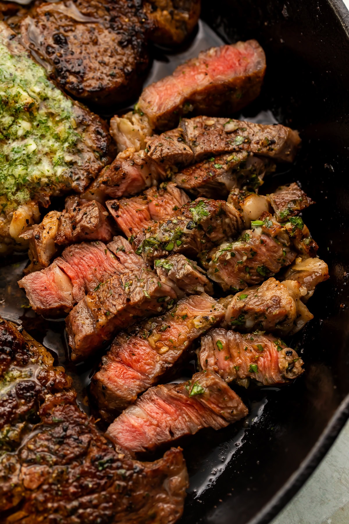 Sliced chuck eye steak in a cast iron skillet with melted parsley herb butter over the top.