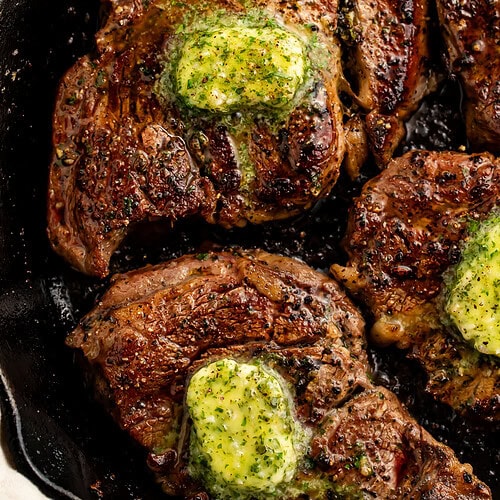Close up of seared chuck eye steaks in a cast iron skillet topped with a garlic herb compound butter.