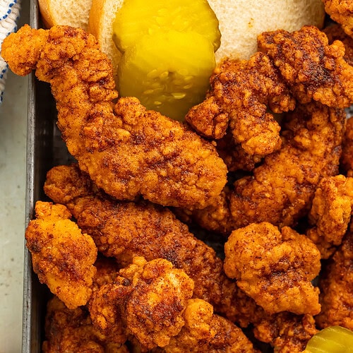 Close up of a tray of Nashville hot chicken tenders with pickle and white bread slices on the side.