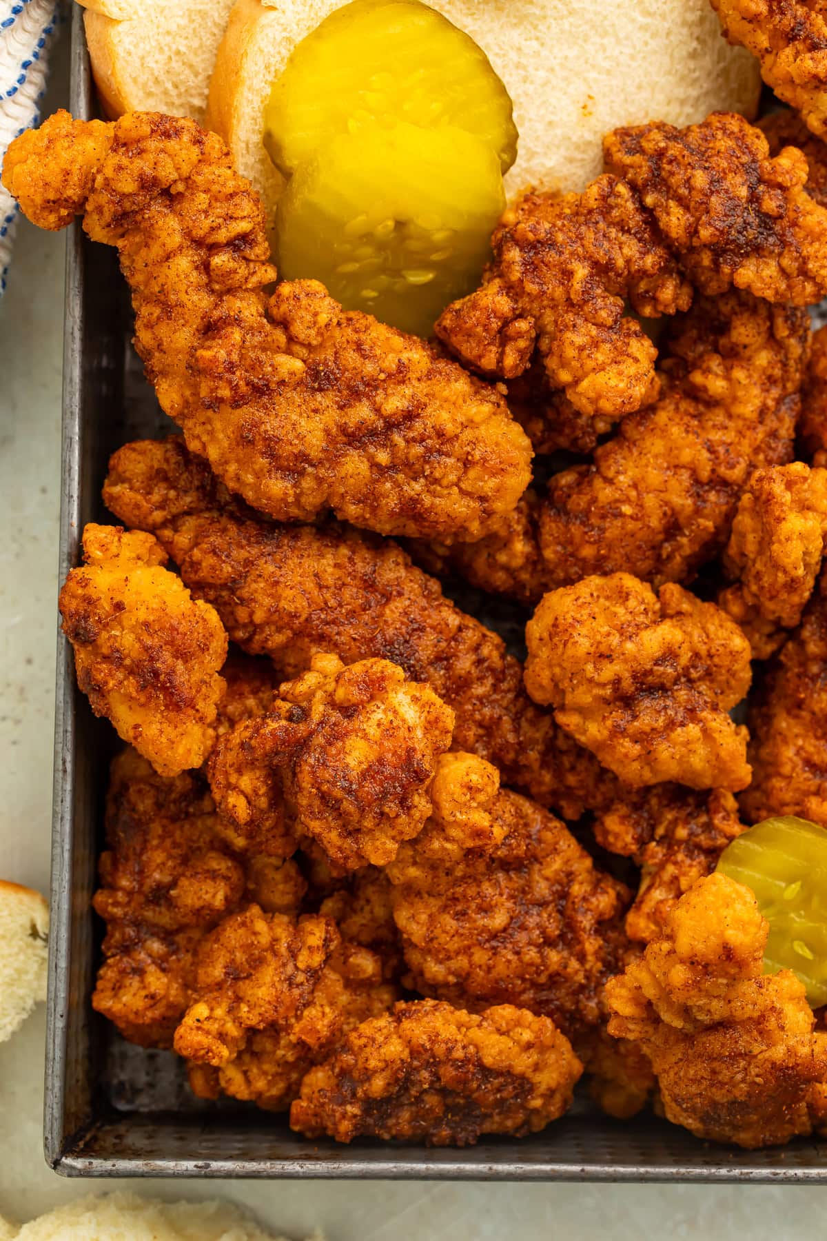 Close up of a tray of Nashville hot chicken tenders with pickle and white bread slices on the side.