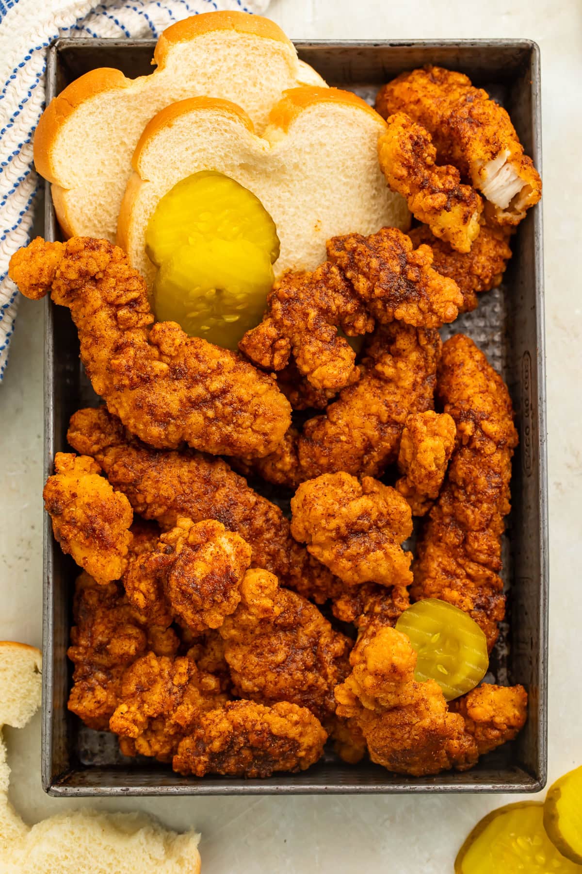 Overhead view of a tray of hot chicken tenders with white bread and sliced pickles.